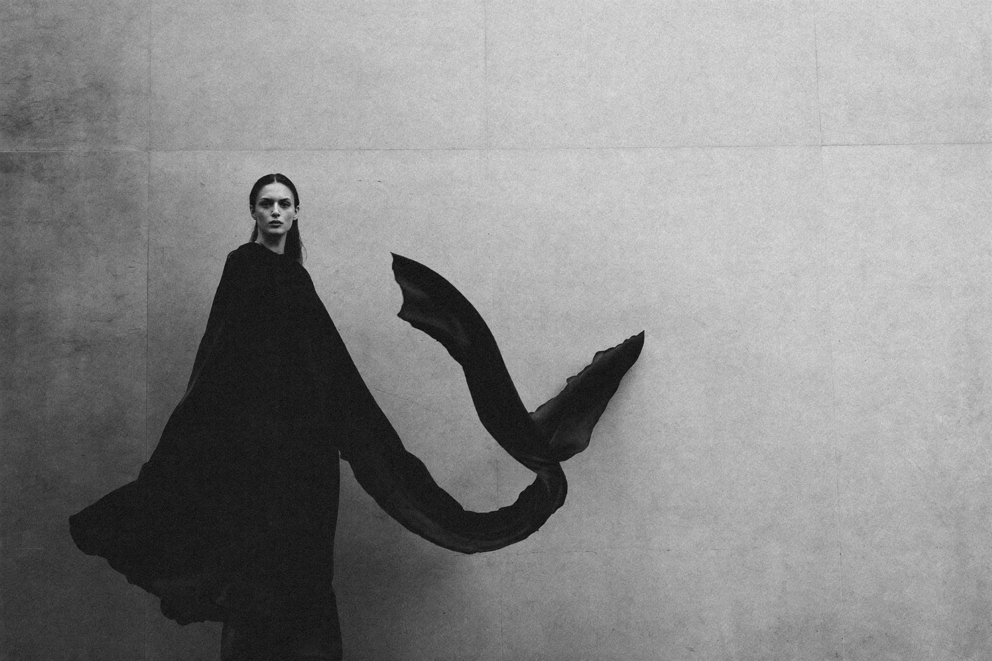 Wide monochrome study of a model in a flowing black draped garment against a pale wall.